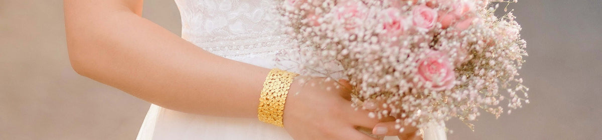 Goldener Armreif an einer Braut mit rosa Brautstrauß und weißem Hochzeitskleid als eleganter Brautschmuck.