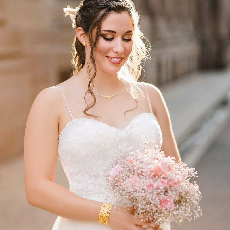 Braut im weißen Hochzeitskleid mit Halskette, Ohrringen und goldenem Armreif sowie rosa Brautstrauß.